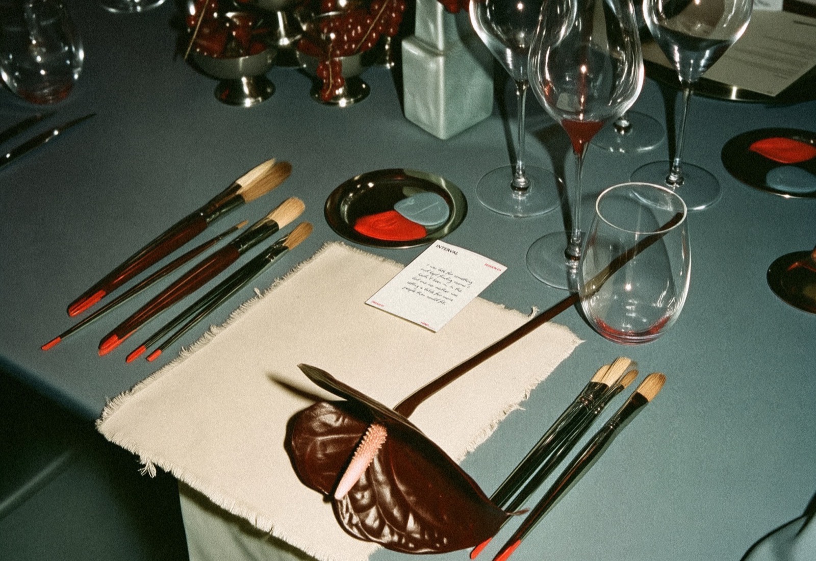 A place setting at the Interval table — brushes, a dream card, an anthurium and wine glasses on blue cloth
