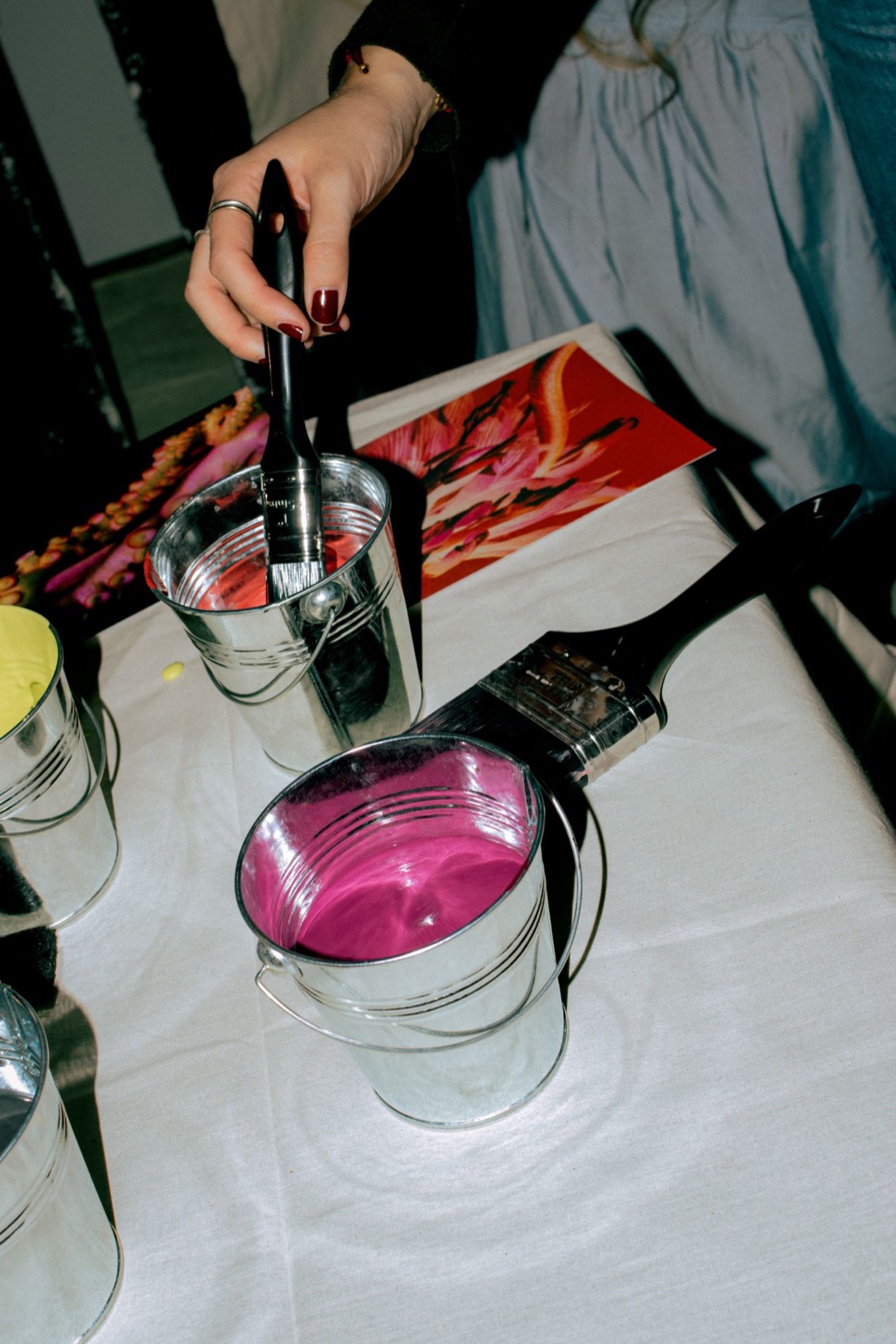 A hand lifting a brush from a pail of magenta paint beside a printed reference image, at a painting evening in London